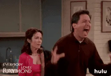 A man joyfully raises his arms and shouts, while a woman in a stylish outfit looks on with a smirk in a living room setting