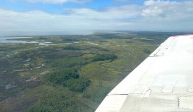 The image shows an aerial view of the Chesapeake Bay, with groves of trees, rivers, a body of water in the distance, and green land. The horizon, a third of the way down the image, separates the blue cloudy sky from the land. On the right of the image is a white wing of a plane.