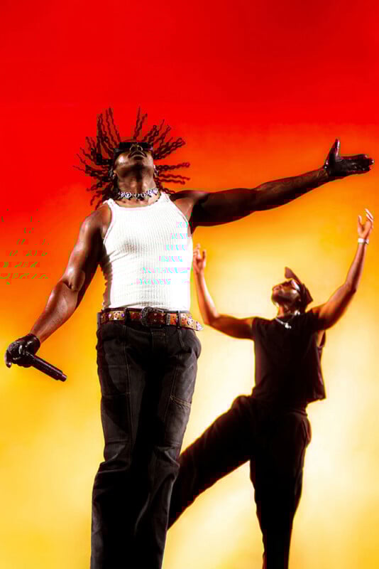 Two performers are on stage against a vibrant red and orange background. The person in front has dreadlocks flying, wearing a white tank top and gloves, holding a microphone with arms wide open. The person in back has arms raised.