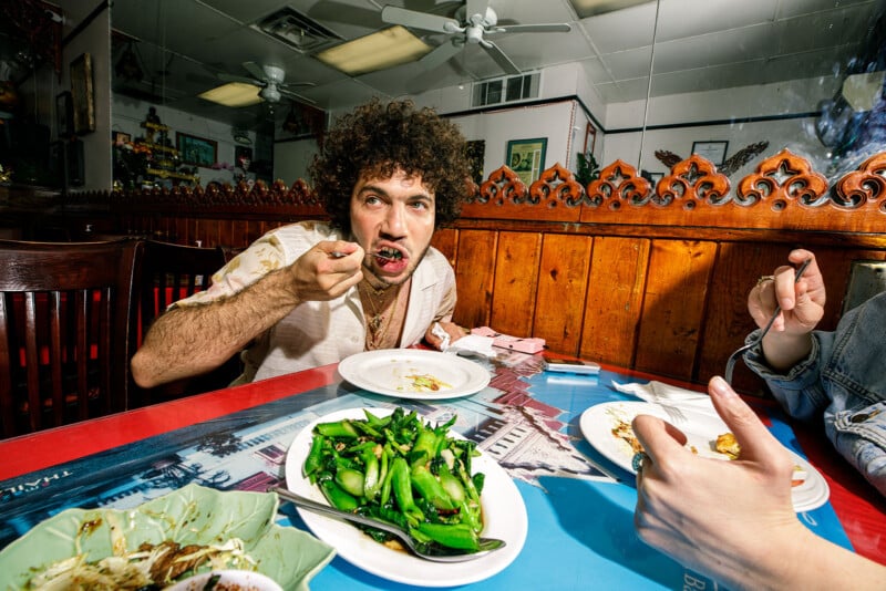 A man with curly hair eats at a restaurant table, holding a fork to his mouth. Green vegetables and plates of food are in front of him. Another person is partially visible, reaching for food. The setting is bright and colorful.