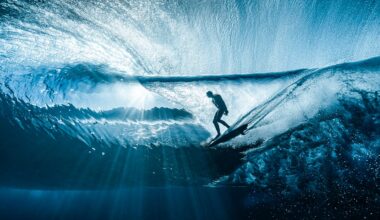A surfer rides inside a translucent blue wave, surrounded by shimmering light rays that filter through the water, creating a dramatic and dynamic underwater perspective.