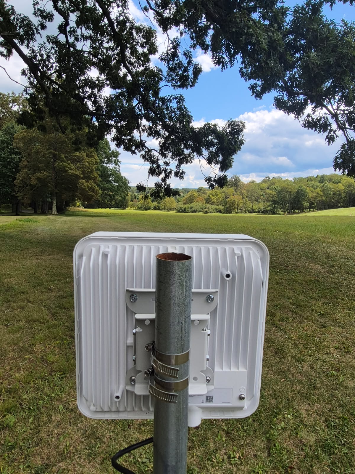 A close-up view of the back of a mounted rectangular wireless antenna fixed to a metal pole with hose clamps. A cable runs from the bottom of the device.