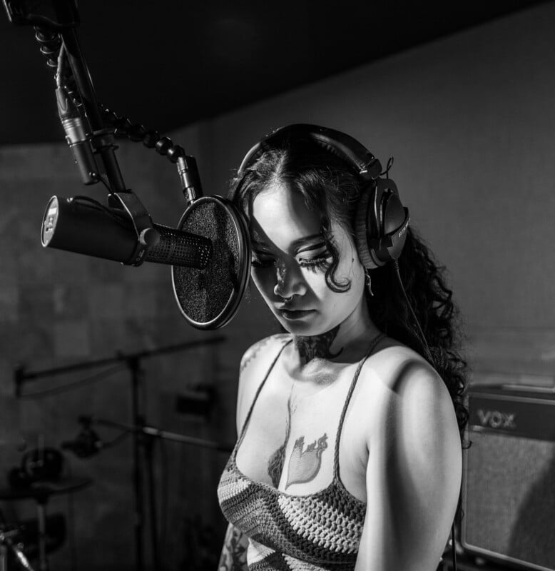 A woman wearing headphones stands in front of a microphone in a recording studio, looking down as she prepares to sing. The image is in black and white.