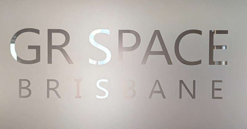 Frosted glass with cut-out letters partially reading "GR SPACE BRISBANE," with the background faintly visible through the transparent text.