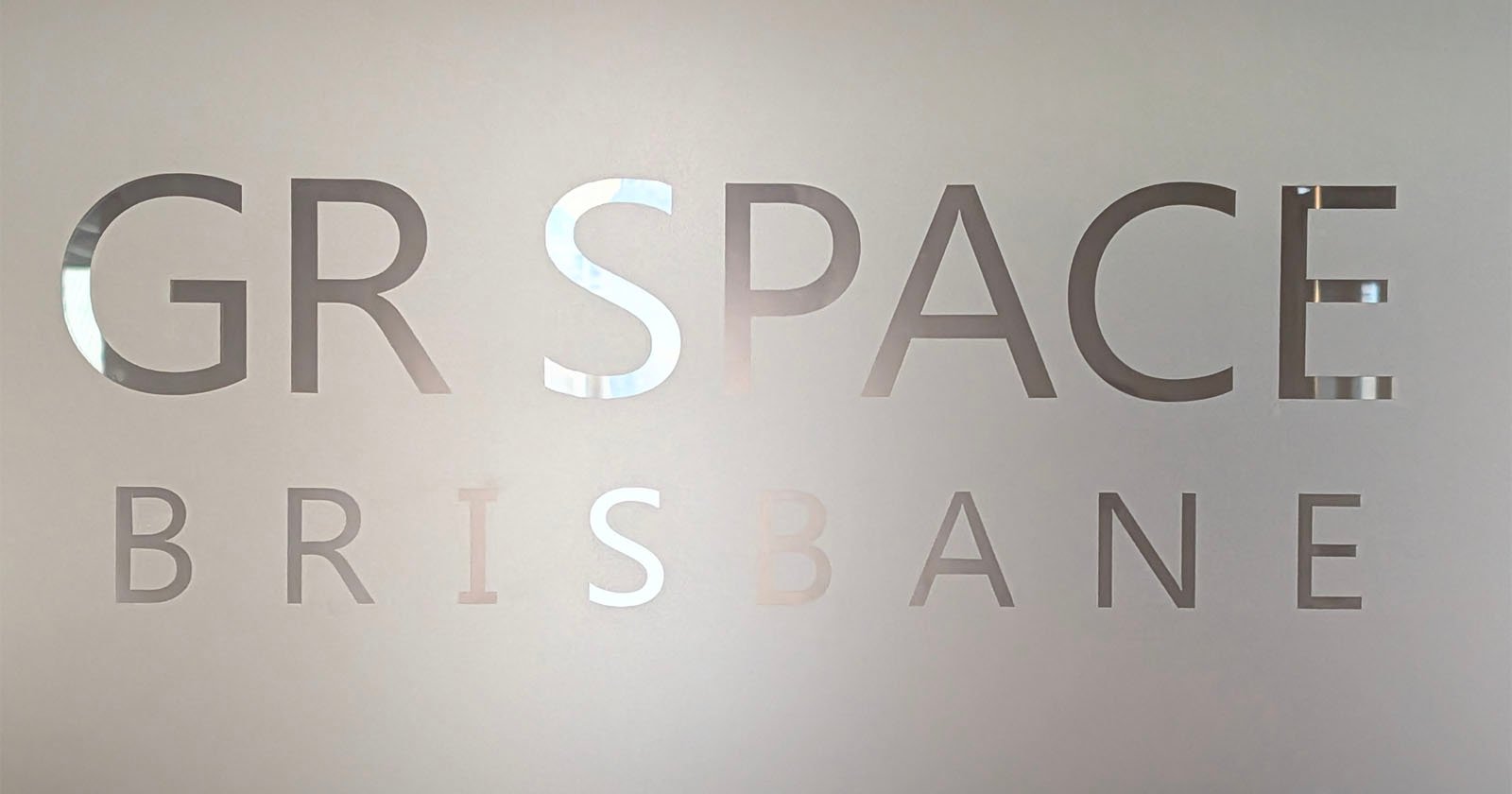 Frosted glass with cut-out letters partially reading "GR SPACE BRISBANE," with the background faintly visible through the transparent text.