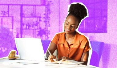 A woman sitting at a desk, writing in a notepad. A laptop is also on the table.
