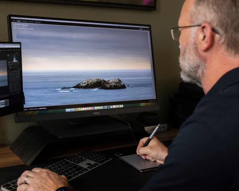 A man uses a stylus and graphics tablet to edit a photo of an ocean scene with a rocky island on a large computer monitor in a home office setting.