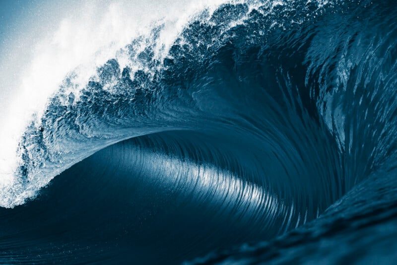 A powerful ocean wave curls and crashes, forming a dramatic barrel with white foam at the crest and deep blue water below.
