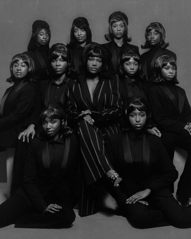 A black-and-white photo of twelve women, all wearing dark suits with similar hairstyles, arranged in three rows. The woman in the center wears a pinstriped suit and sits confidently, while the others surround her closely.