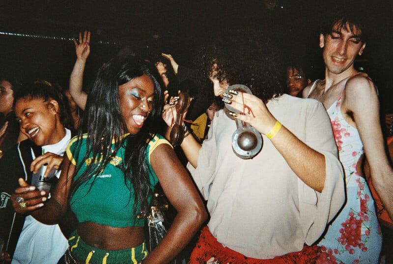 A lively group of people dance closely together at a crowded party. They are smiling, laughing, and appear to be enjoying the music, with drinks in hand and colorful outfits. The atmosphere is joyful and energetic.