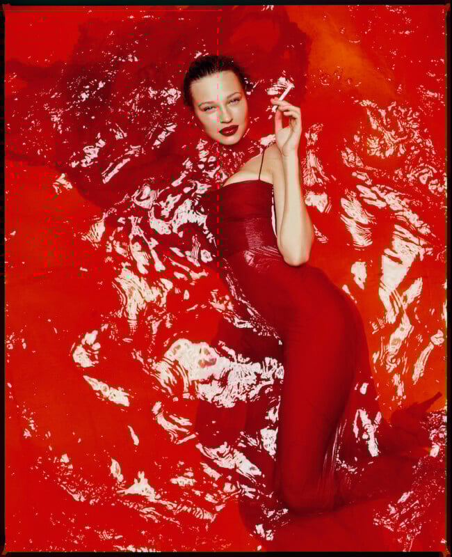 A woman in a form-fitting red dress lies in vibrant red liquid, holding a cigarette, with slicked-back hair and bold red lipstick. The fluid’s ripples create dramatic reflections around her.