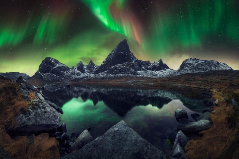 A vivid aurora borealis glows green and pink above sharp, snow-dusted mountains reflected in a calm lake, with rocky terrain and grasses in the foreground under a starry night sky.