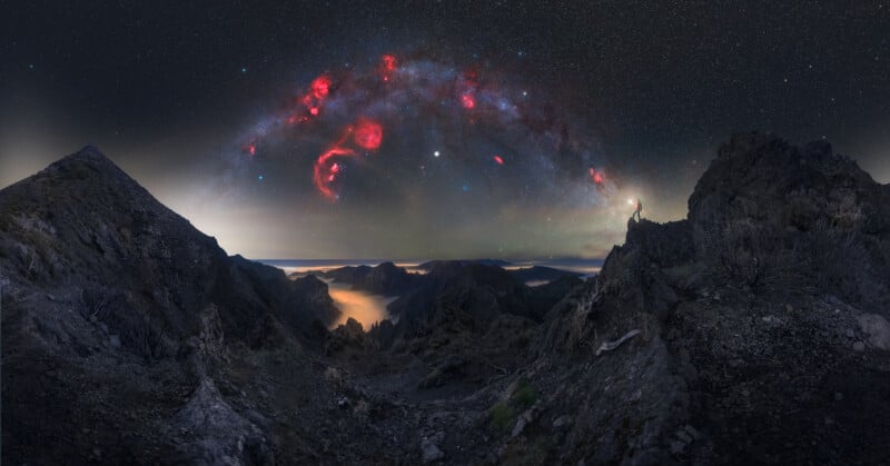 A person stands on a rocky mountain landscape at night, shining a light toward a dramatic sky filled with bright stars and vibrant red nebulas arched over the horizon.
