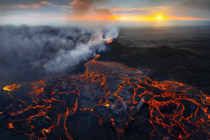 Aerial view of an erupting volcano at sunset, with glowing lava flows, cracked black ground, and smoke rising into the sky. The horizon is lit by the setting sun.
