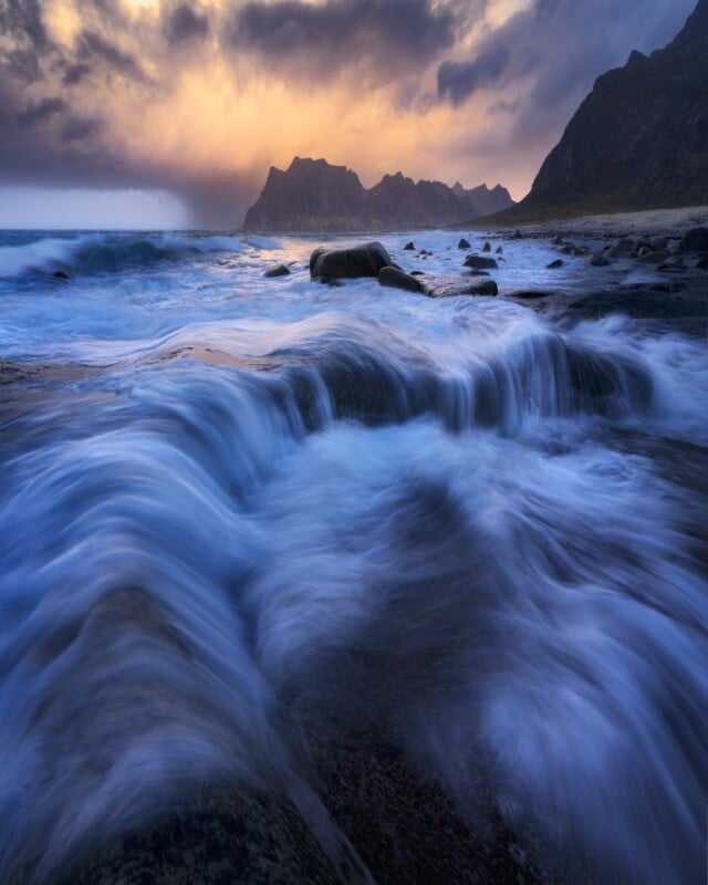 Waves rush over rocks on a dramatic coastline at sunset, with dark mountains silhouetted against a colorful sky of orange, purple, and blue clouds.