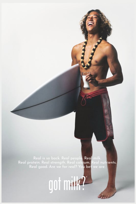 A smiling young man holding a surfboard and a bottle of milk stands shirtless, wearing board shorts and a beaded necklace. The text at the bottom reads: “got milk?” with additional promotional phrases above.