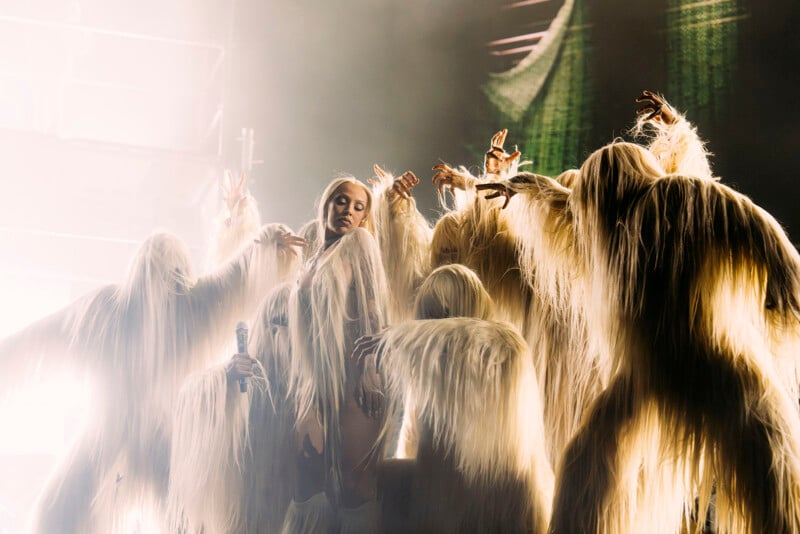 A performer in a dramatic, long, white, shaggy costume stands surrounded by dancers in similar outfits, all reaching towards the center under theatrical lighting on stage.