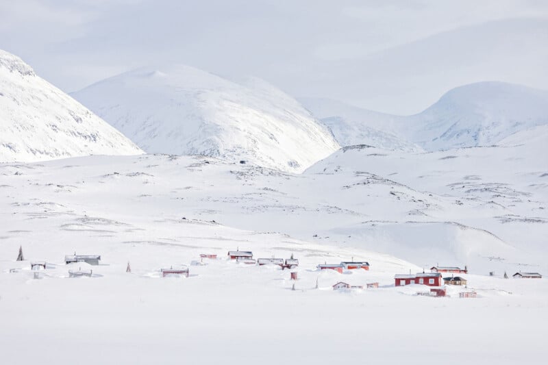 Small houses with red and white roofs are scattered across a snowy, mountainous landscape under a bright, overcast sky. Snow-covered peaks rise in the background, creating a serene, wintry scene.