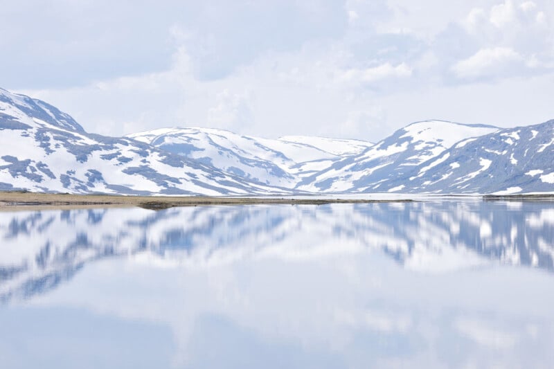 A calm lake reflects snow-covered mountains under a cloudy sky, creating a serene and symmetrical landscape with soft, cool tones.