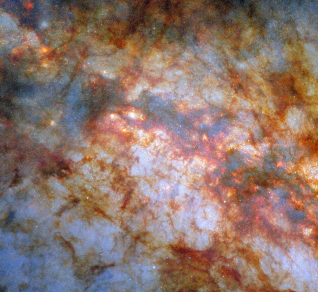 A close-in view of the center of galaxy M82. Bright, bluish light radiating from the center is due to stars actively forming there. A thick lane of gas, black in the center and red around the edges, crosses the center and blocks much of the light. Thinner strands and clumps of reddish dust cover much of the rest of the view.