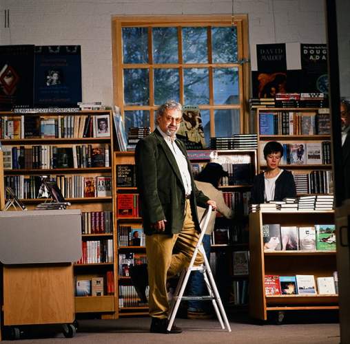 A man wearing amber glasses and a green suit jacket leans against a foot ladder in front of bookshelves where people are browsing.