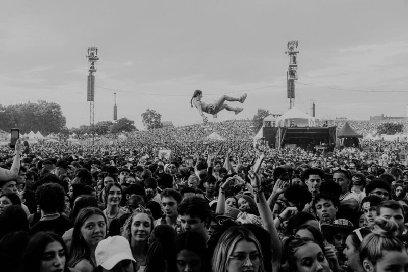 A large crowd at an outdoor music festival with a person crowd surfing above the audience. The photo is in black and white, and the stage and towers are visible in the background.