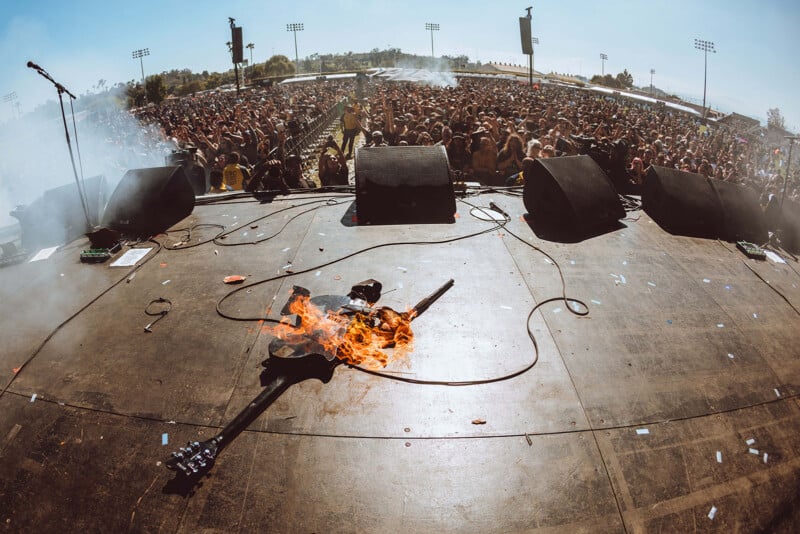 A burning electric guitar lies on an outdoor concert stage, with a large cheering crowd and stadium lights visible in the background under a bright sky.