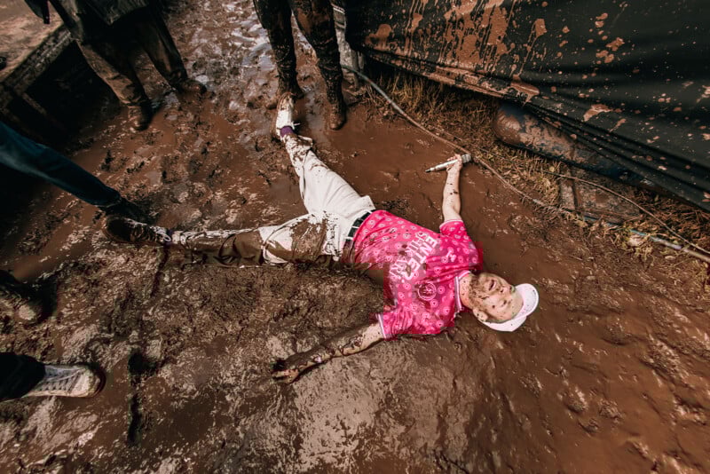 A person wearing a pink shirt, white cap, and white pants lies on their back in thick mud, arms and legs spread out, with mud covering much of their body. People stand nearby on the muddy ground.