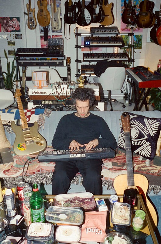 A person sits on a couch playing a Casio keyboard, surrounded by guitars, music equipment, food containers, and drinks, in a cluttered, creative studio space.