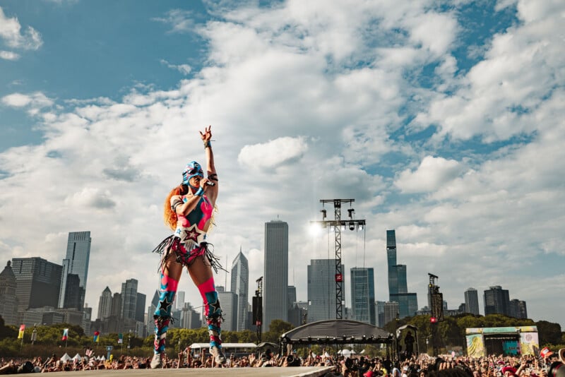 A performer in a colorful costume poses on an outdoor stage with one arm raised, in front of a large crowd and a city skyline under a partly cloudy sky.