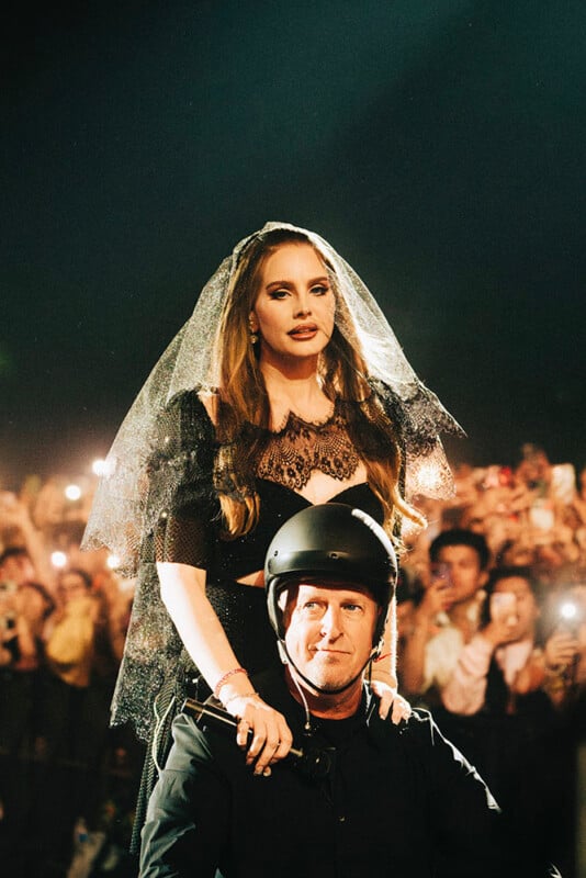 A woman in a black lace dress and veil stands behind a man in a black helmet, with her hands on his shoulders. They are in front of a crowd holding up phones and lights at what appears to be a concert.