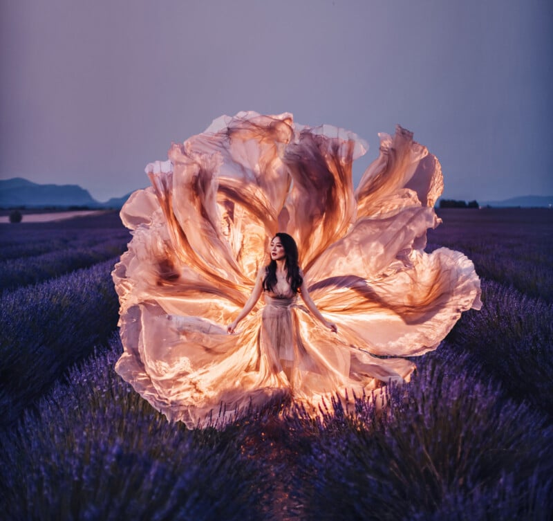 A woman stands in a lavender field at dusk, surrounded by a flowing, illuminated dress that fans out dramatically around her, creating a flower-like shape against the purple landscape.