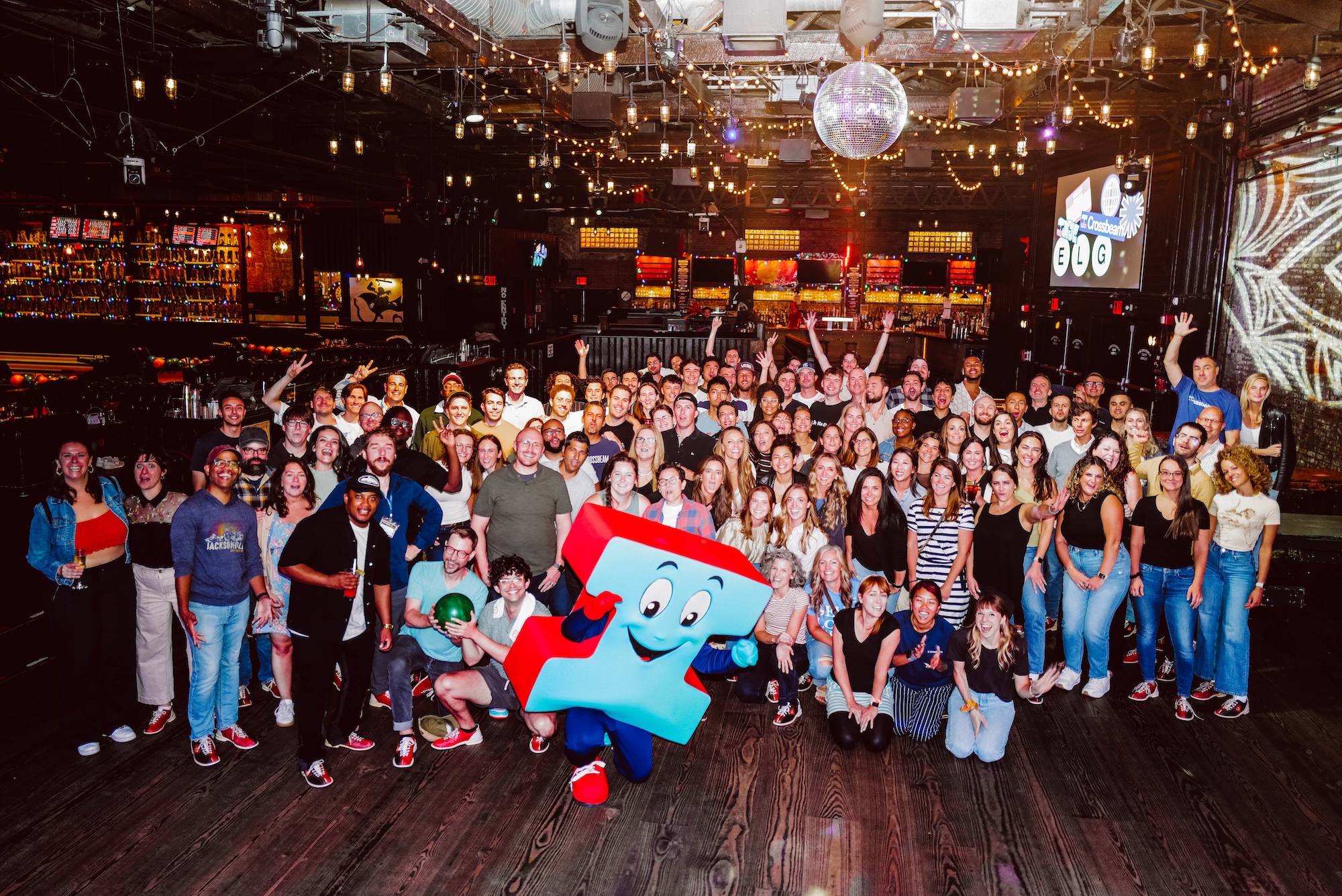 A large group of people pose for a photo indoors with a puzzle piece mascot in front; colorful lights and bar area visible in the background.