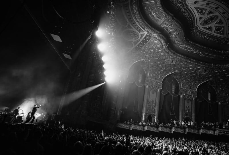 A black and white photo of a band performing on stage in a grand, ornate theater filled with a large, cheering audience; dramatic lighting highlights the intricate ceiling and enthusiastic crowd.