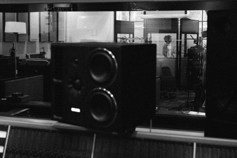 Black and white photo of a music studio with a large speaker in the foreground and a person standing in a soundproof booth singing or speaking into a microphone in the background.