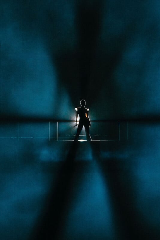 A silhouette of a person stands with legs apart and hands on hips, backlit by bright light, creating dramatic shadows and a blue, smoky atmosphere.