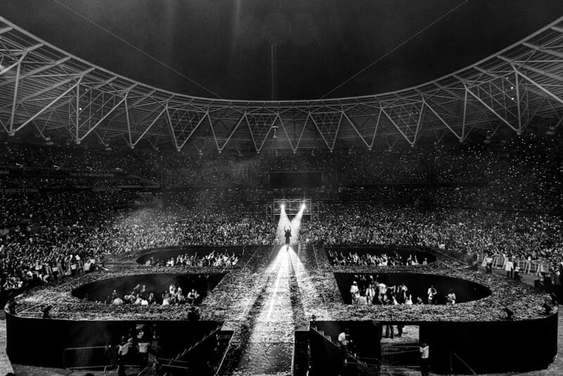A black and white photo of a large, circular stadium filled with a crowd at a concert. Spotlights shine on a performer standing at the center of a circular stage, surrounded by confetti and cheering fans.