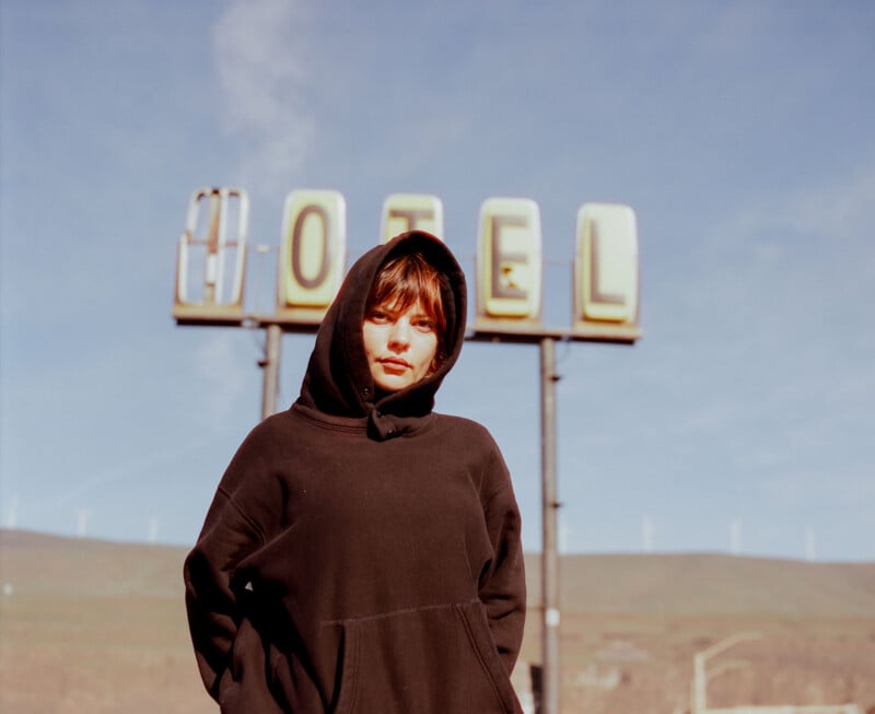 A person wearing a black hoodie stands in front of a vintage "HOTEL" sign outdoors, with a blue sky and hills in the background.