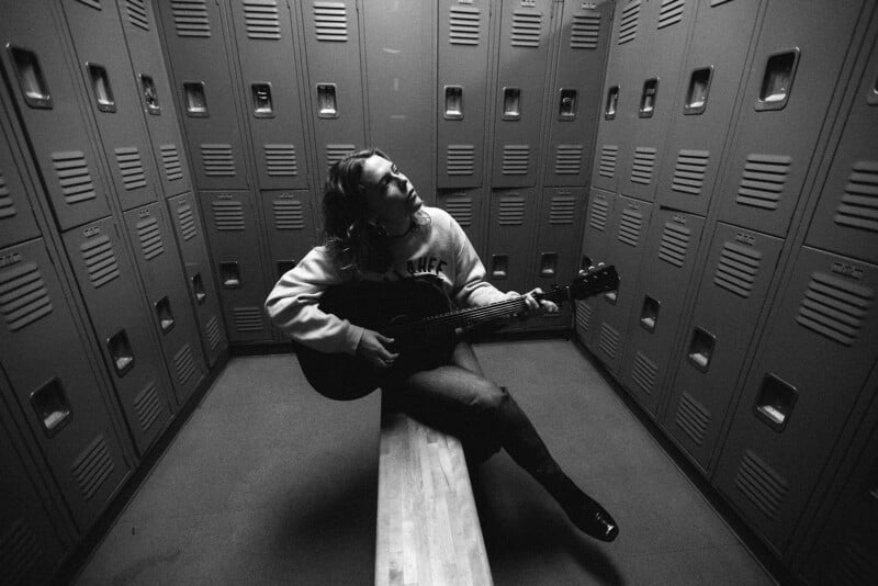 A person sits on a bench in a locker room, playing an acoustic guitar. They are surrounded by rows of lockers and appear to be lost in thought. The image is black and white.