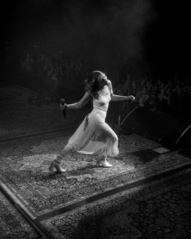 A woman in a flowing dress holds a microphone and dances energetically on a stage, illuminated by spotlight, with an audience cheering in the background. The image is in black and white.