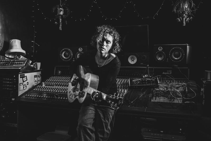 A person with curly hair sits in a dimly lit music studio, playing an acoustic guitar in front of a large audio mixing console, surrounded by speakers, cables, and studio equipment.