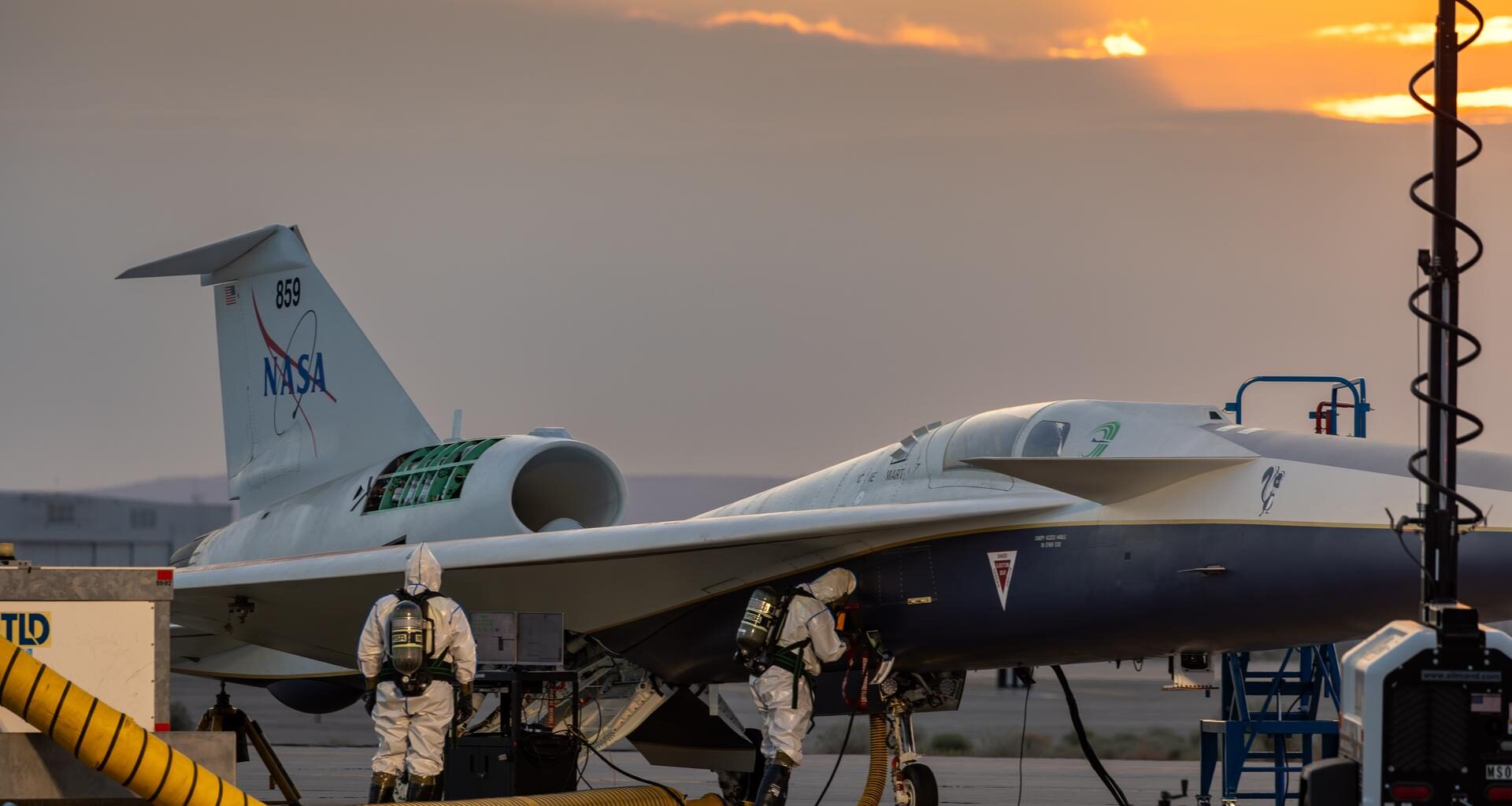 Two maintainers in white protective suits, air tanks, masks, rubber gloves, and boots check NASA’s X-59 hydrazine system during a hydrazine safety check at U.S. Air Force Plant 42 in Palmdale, California. The sky is slightly overcast, with some sunlight rays beginning to peek out from behind the clouds. The aircraft has a panel open, a ladder positioned on the right side of its fuselage, and air ducts extending from the aircraft into a service cart.