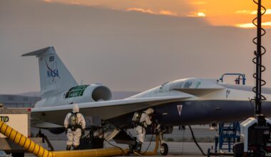 Two maintainers in white protective suits, air tanks, masks, rubber gloves, and boots check NASA’s X-59 hydrazine system during a hydrazine safety check at U.S. Air Force Plant 42 in Palmdale, California. The sky is slightly overcast, with some sunlight rays beginning to peek out from behind the clouds. The aircraft has a panel open, a ladder positioned on the right side of its fuselage, and air ducts extending from the aircraft into a service cart.