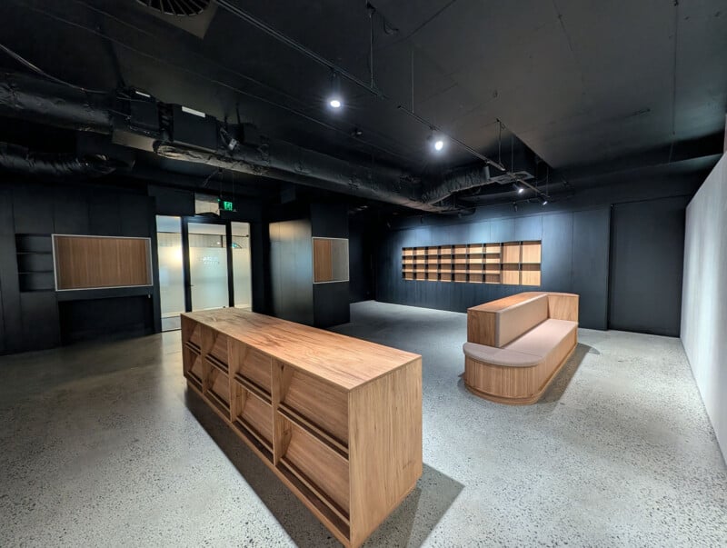 Modern, minimalist interior with black walls and ceiling, wooden shelving units, a central wooden island, and a wooden bench with a cushioned seat on polished concrete flooring. Glass doors are visible at the back.