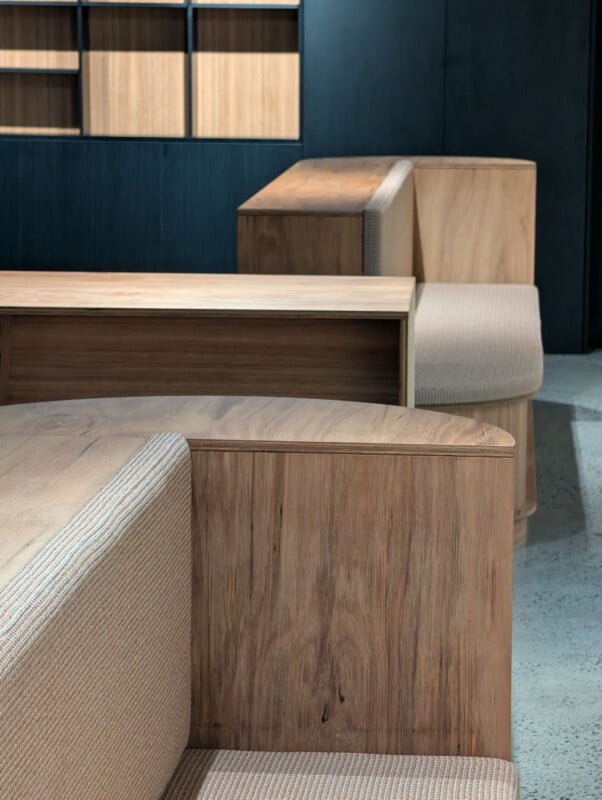 Modern wooden benches with light beige upholstered seating are arranged in a minimalist interior with concrete flooring and wooden shelving against a dark wall in the background.