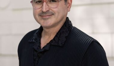 The person is wearing a black, textured outfit with pleated fabric and a detailed collar design. The background consists of light-colored brickwork.