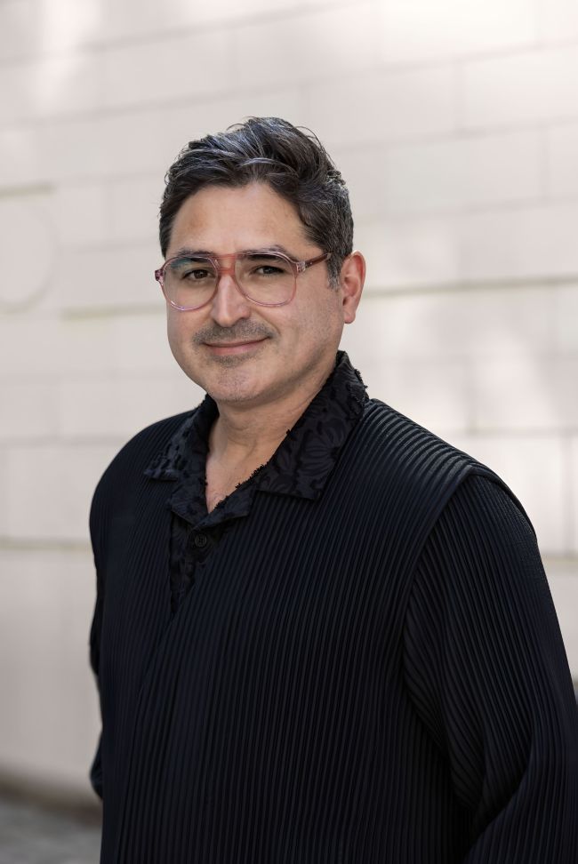 The person is wearing a black, textured outfit with pleated fabric and a detailed collar design. The background consists of light-colored brickwork.