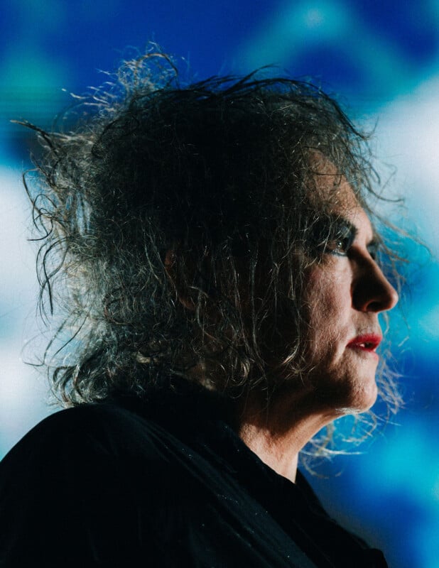 A person with messy, voluminous gray hair and dramatic makeup is shown in profile against a blue and white blurred background, wearing a dark shirt.