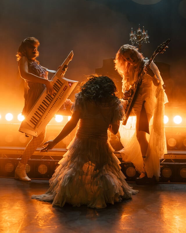 Three women in elegant, vintage-style dresses perform on stage with bright orange backlighting; one plays a keytar, one plays an electric guitar, and one kneels at the center, facing away from the camera.