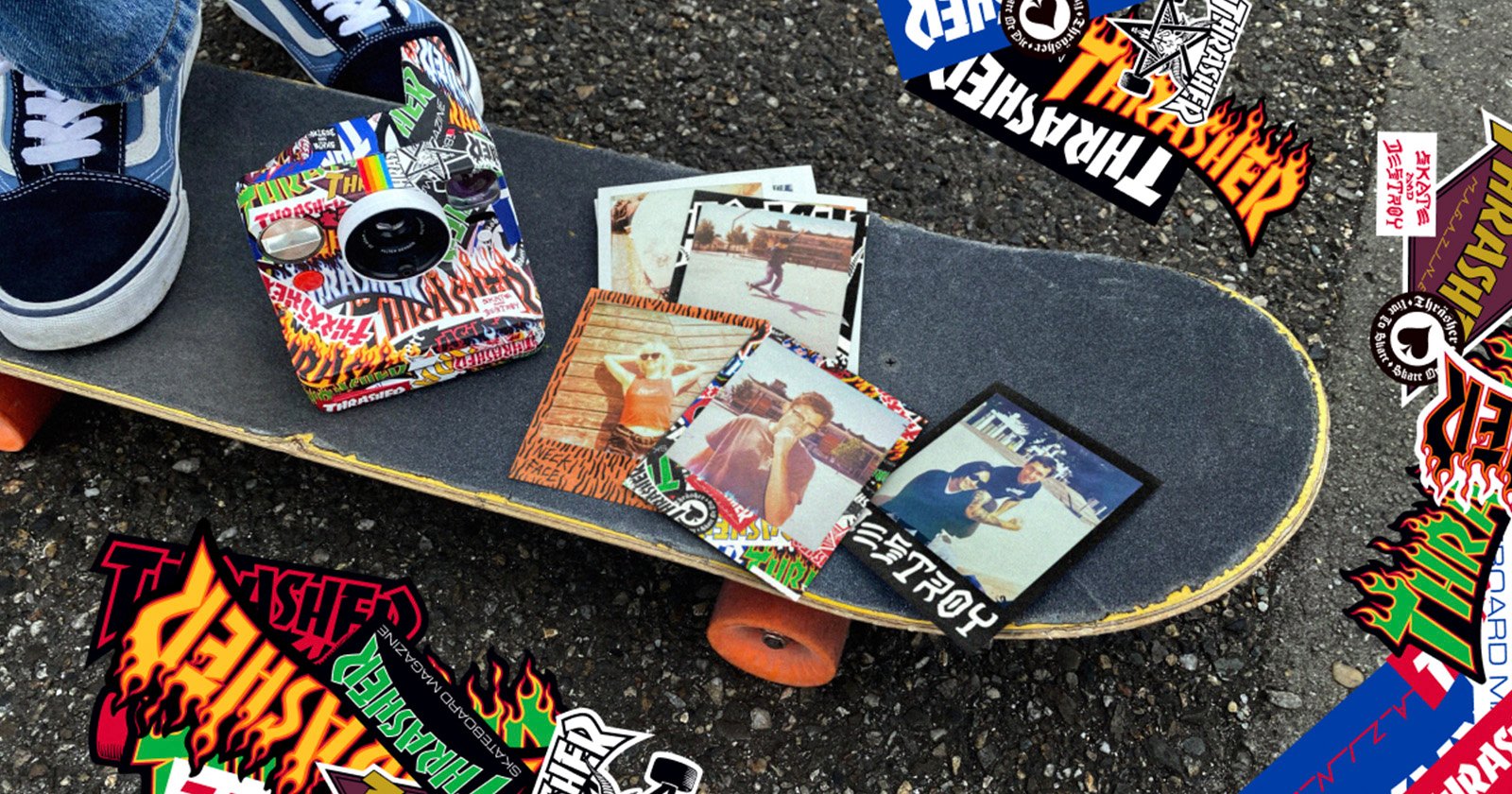A skateboard with orange wheels rests on asphalt, covered in colorful "Thrasher" stickers. Several retro-style photos and more stickers are scattered on the skateboard. A person’s feet in blue sneakers are visible nearby.